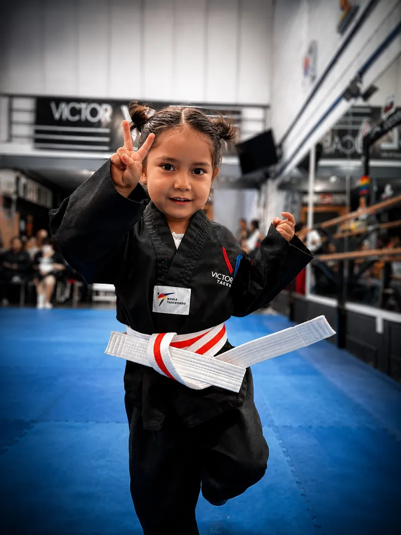 Niña pequeña con dobok negro y cinta blanca sonriendo durante su clase de Taekwondo para preescolares.