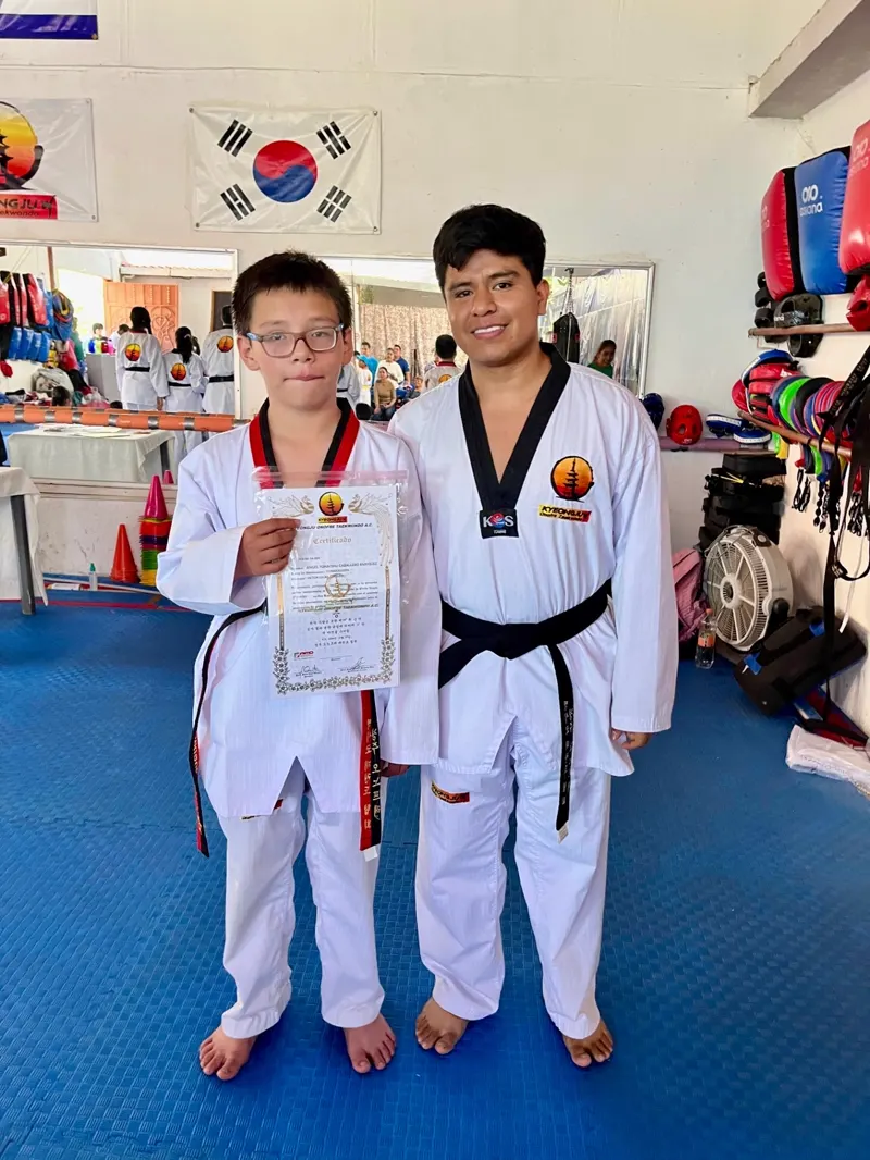 Alumno recibiendo su certificado de grado junto a su instructor frente a la bandera de Corea en el dojang de Kyeongju.
