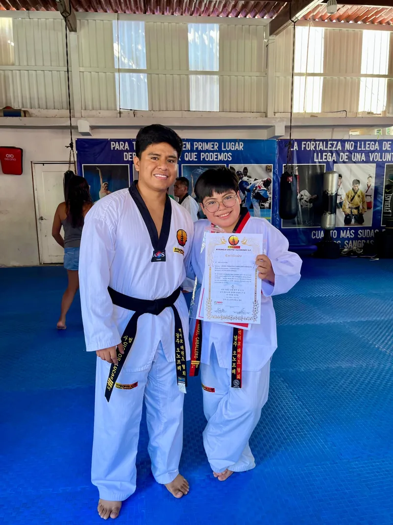 Alumno de Kyeongju Onofre Taekwondo mostrando su certificado de grado acompañado por su maestro en el dojang.